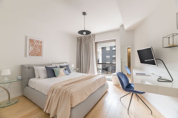A modern hotel room with a bed, a desk, a chair, a TV, and a window with curtains. - Elegant 2BR Apartment in Kolonaki (Athens)