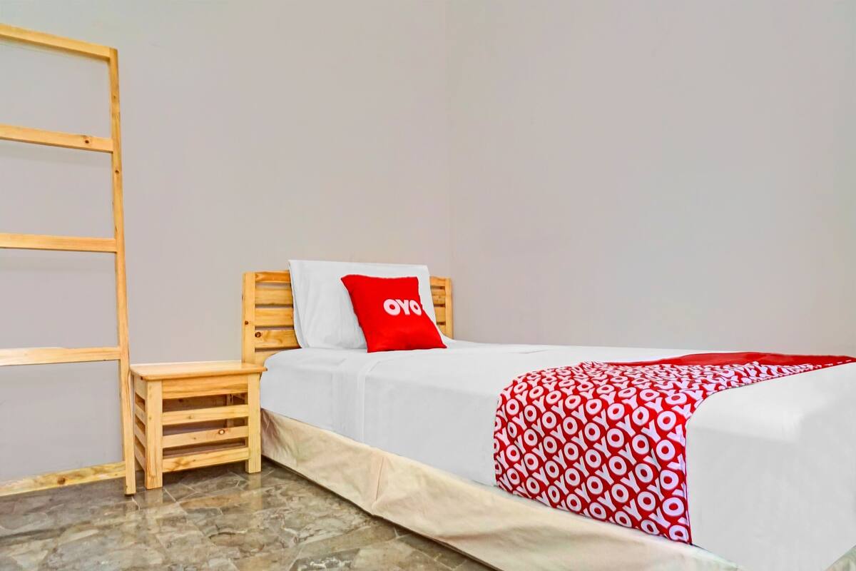 A single bed with a white mattress and a red and white patterned blanket. There is a small wooden stool beside the bed.
