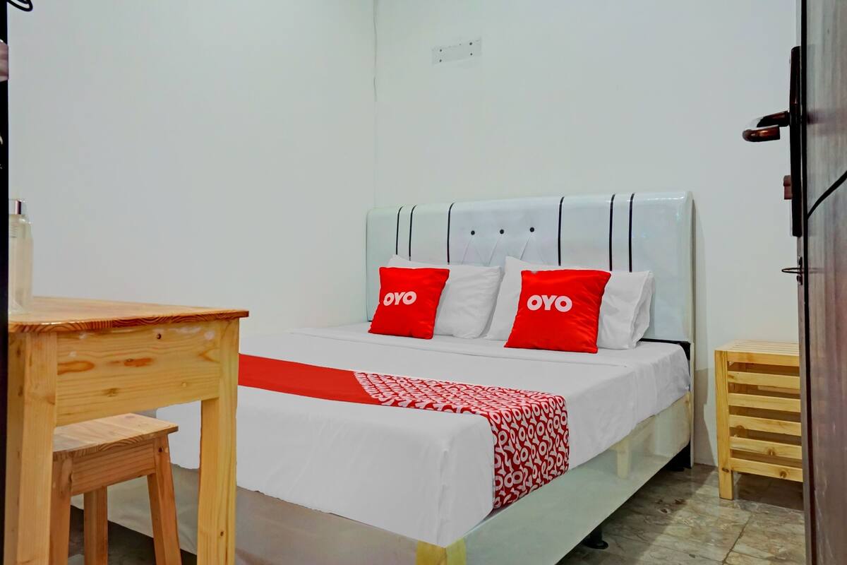 A single bed with a white and red bedspread, two red pillows with the word "OYO" on them, and a wooden nightstand beside the bed.