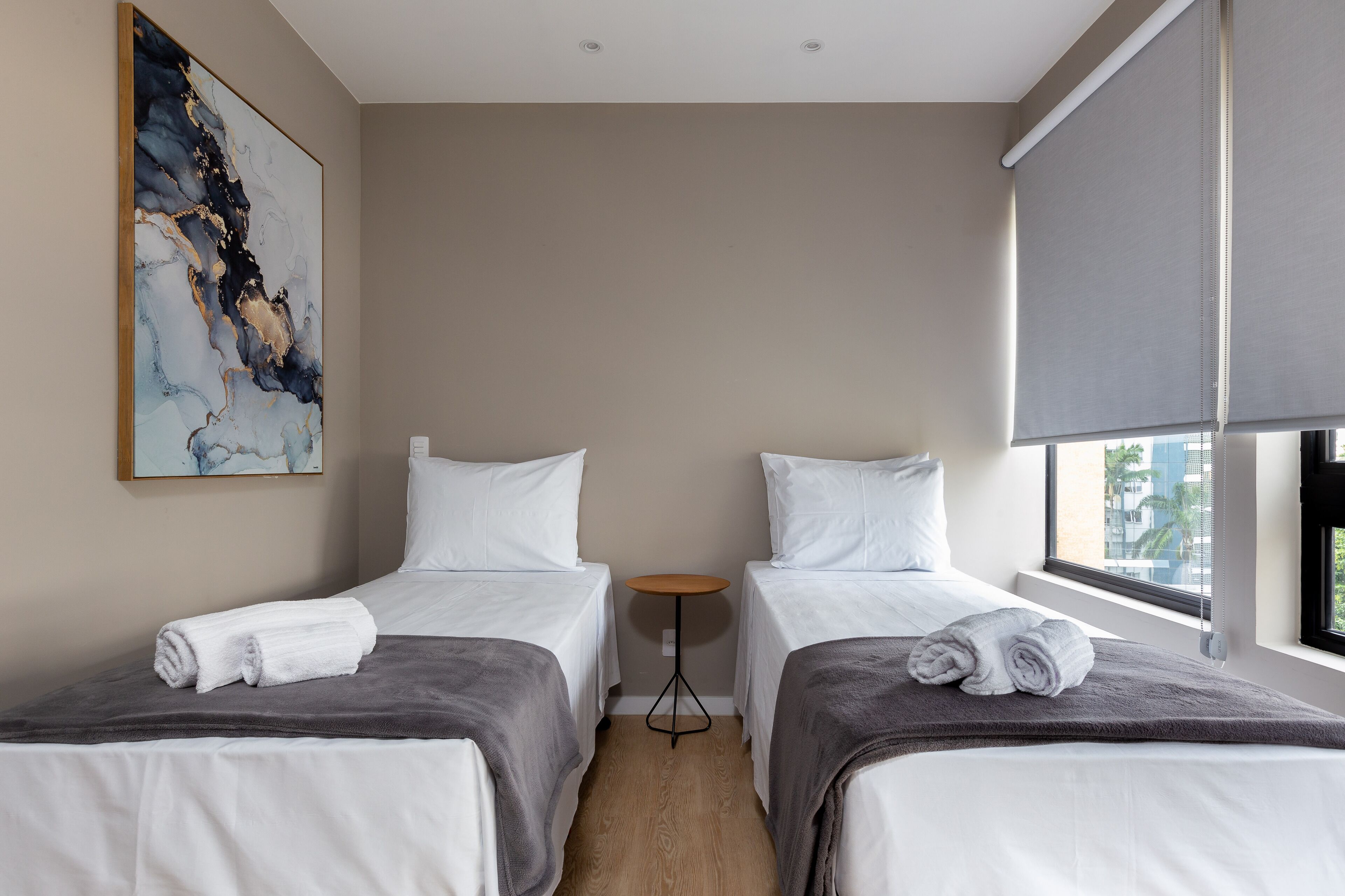 A hotel room with two beds, a small round table, a painting on the wall, and a window with a view of buildings.