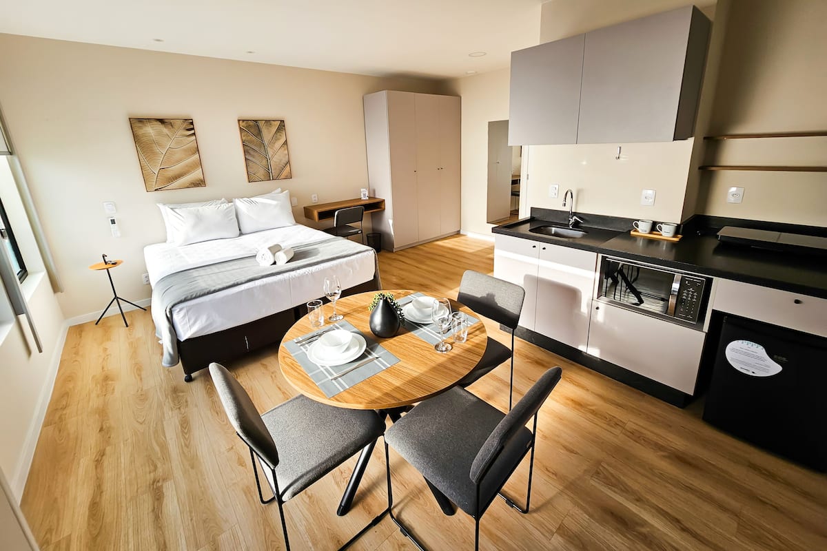 A modern hotel room with a bed, a small round table with chairs, a kitchenette, and a desk.