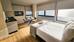 A hotel room with a bed, a desk with a chair, a TV mounted on the wall, and a window with blinds. - Bewiki (Florianópolis)