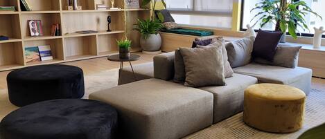 A modern living room with a sectional sofa, ottomans, a bookshelf, and potted plants.