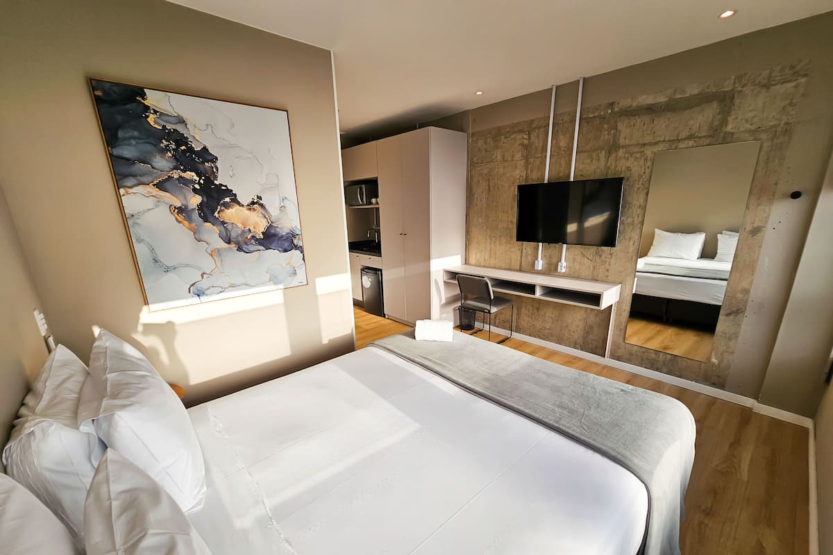 A modern hotel room with a large bed, a desk, a TV, and a kitchenette.