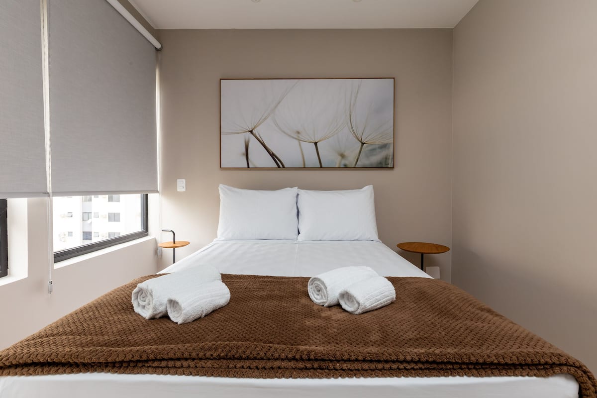 A hotel room with a bed, two bedside tables, a window with blinds, and a wall-mounted artwork.
