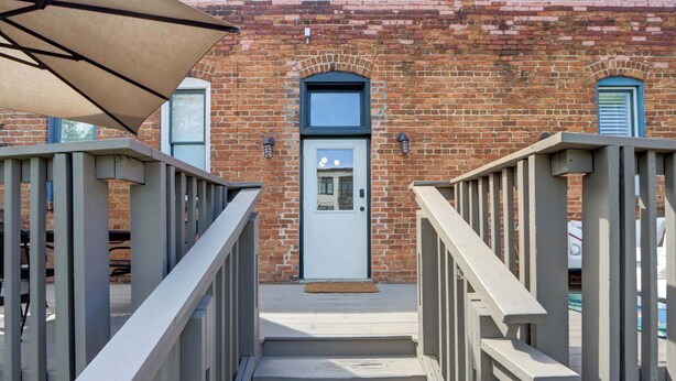 A staircase leading to a door with a small porch area, a brick building with a window and a door, and a metal railing on the left. - Carolina Gold @ The Huntingdon Lofts Collection (Savannah)