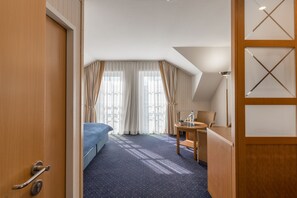 A hotel room with a bed, a desk, and a chair. There is a window with curtains and a view of the outside. - Strandhotel Seestern (Ostseebad Baabe)