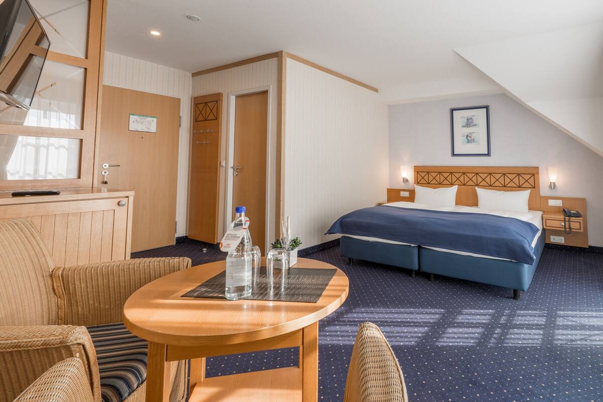 Habitación de hotel con cama, una mesa de madera, una silla y una mesita con botellas y vasos.