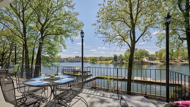 A deck with outdoor seating overlooking a lake, surrounded by trees and a view of buildings in the distance. - Family & Friends Paradise on the lake (Rockwell)