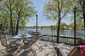 Terraza con asientos al aire libre que da a un lago, rodeada de árboles y con vistas a edificios en la distancia.
