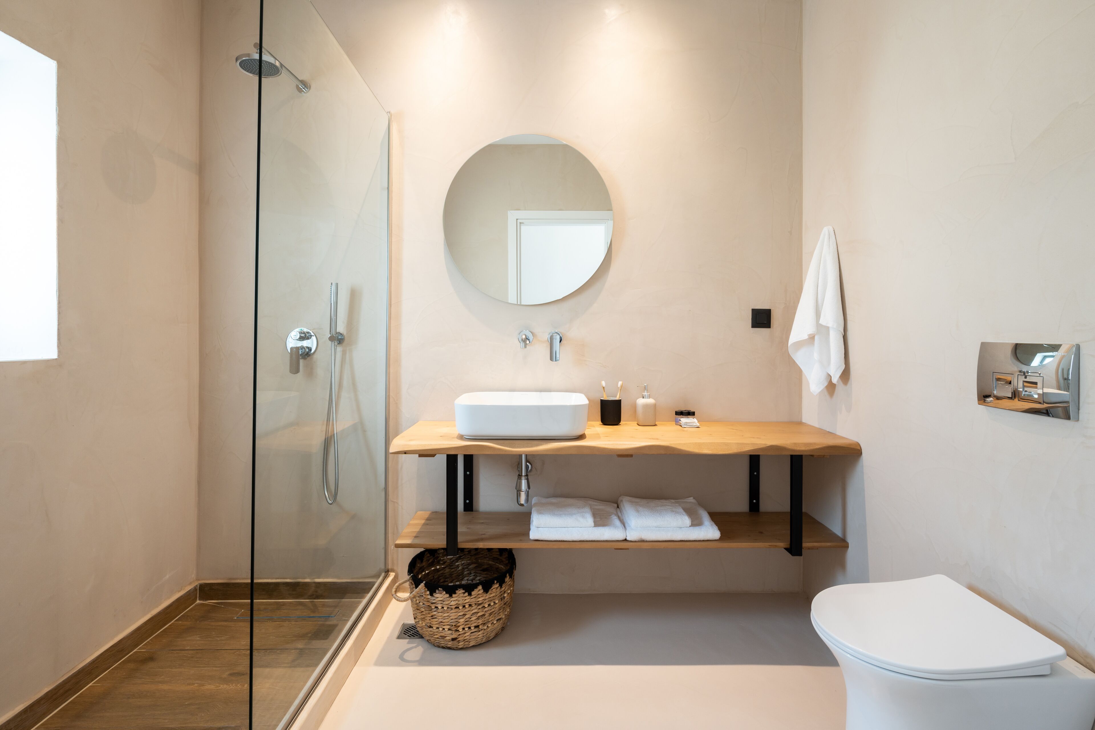 A modern bathroom with a glass shower enclosure, a round mirror, a wooden vanity, and a white toilet.