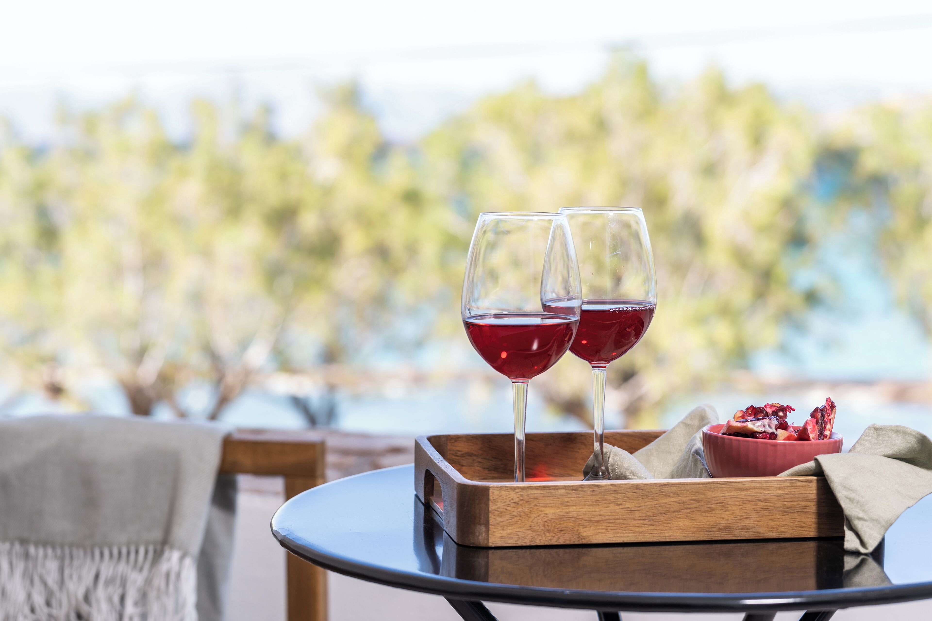 Two wine glasses on a tray with a bowl of food, set on a table with a view of trees and water.
