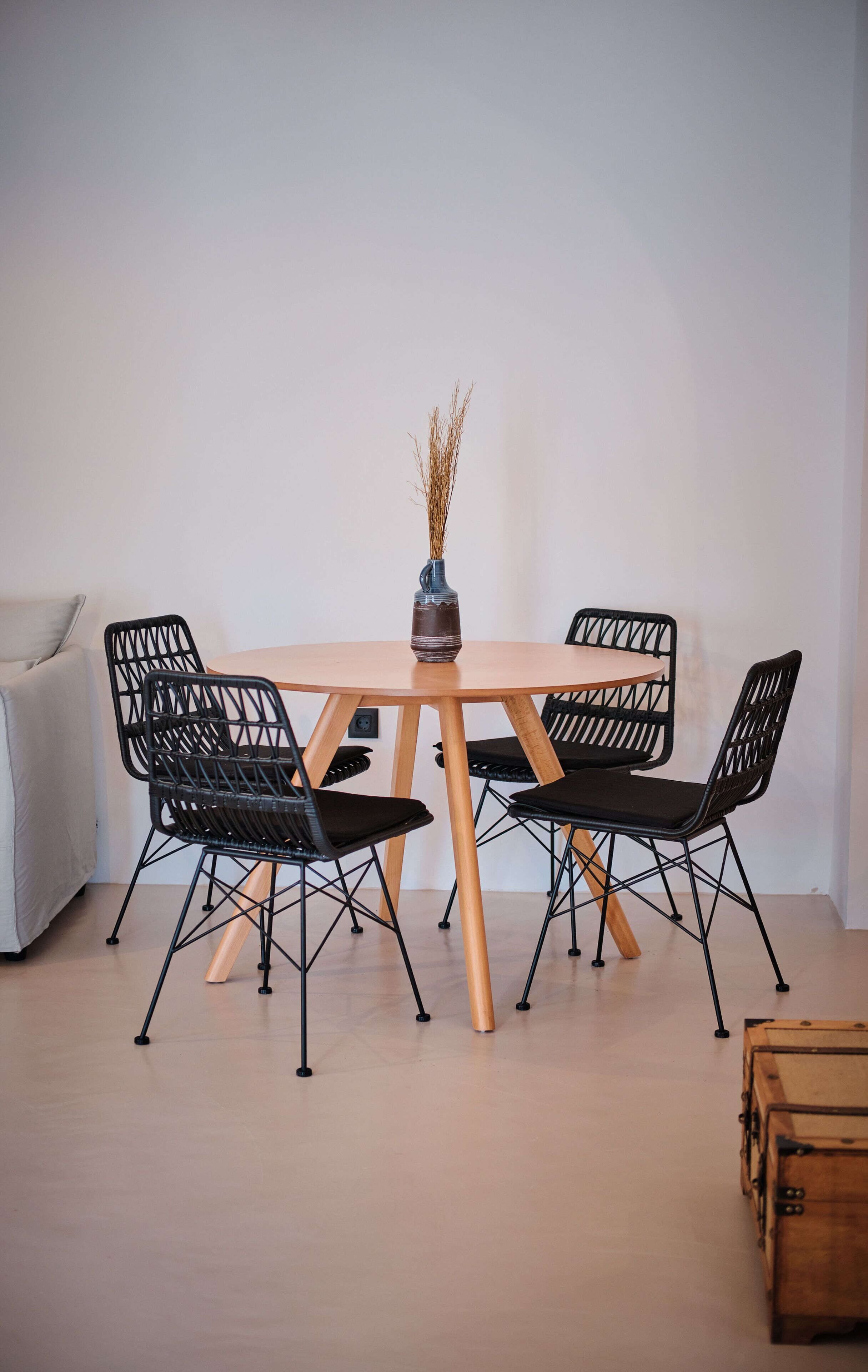 A dining area with a round wooden table, four black chairs, a vase with dried plants, and a wooden chest.