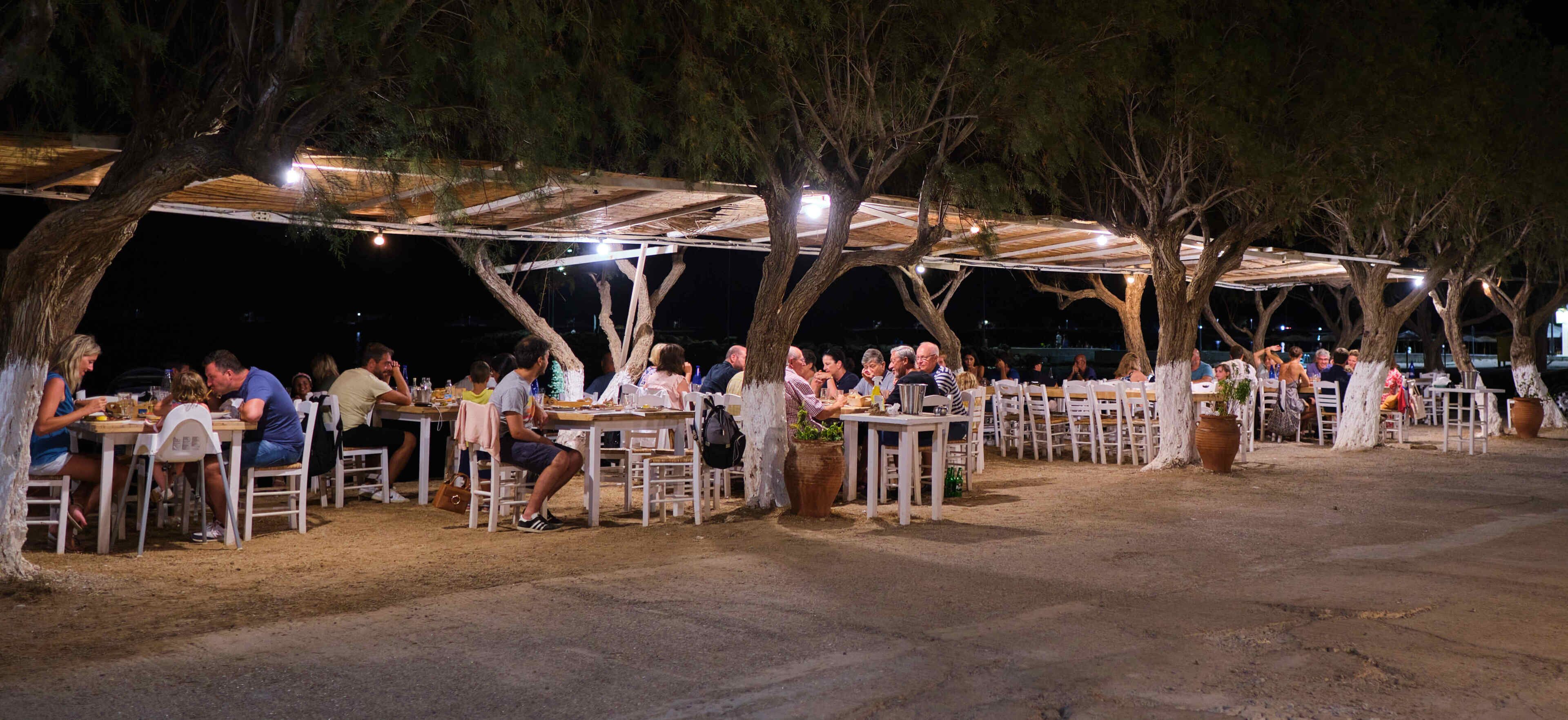 An outdoor dining area with tables and chairs under a covered structure, surrounded by trees.