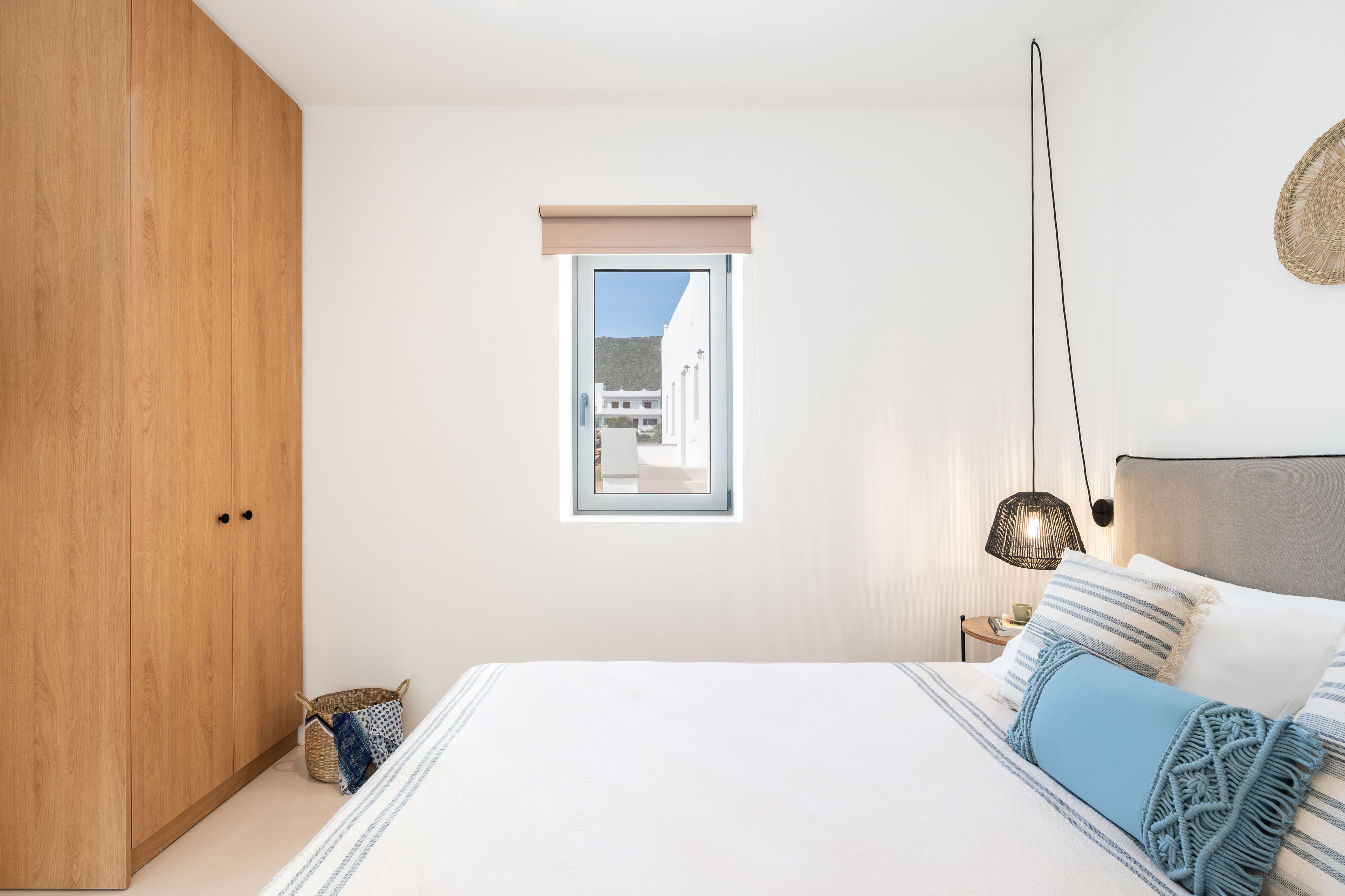 Une chambre avec une grande armoire en bois, une petite fenêtre donnant sur des immeubles, une table de chevet avec une lampe, et un lit avec une literie à rayures et un plaid bleu.