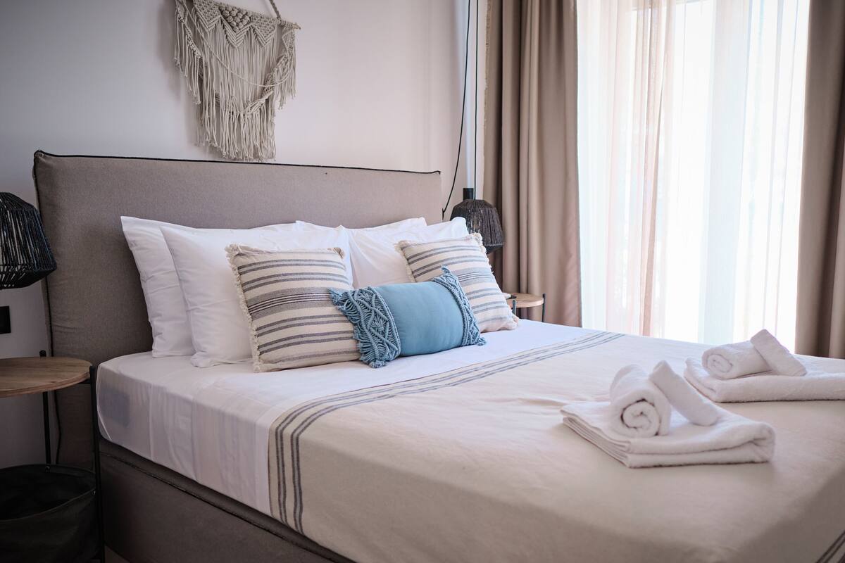 A neatly made bed with white and striped pillows, a blue throw, and rolled towels on a bed in a room with a stone wall and a macrame wall hanging.