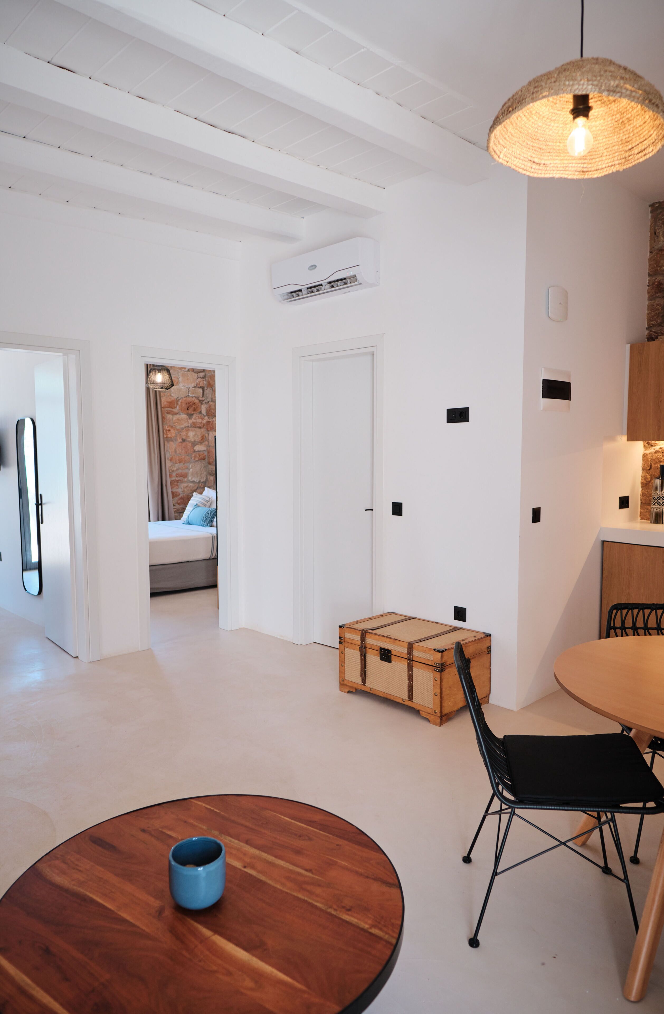 Una habitación moderna y luminosa con una mesa de madera, una taza azul, una silla negra, una maleta y una cama con pared de piedra y cortinas.