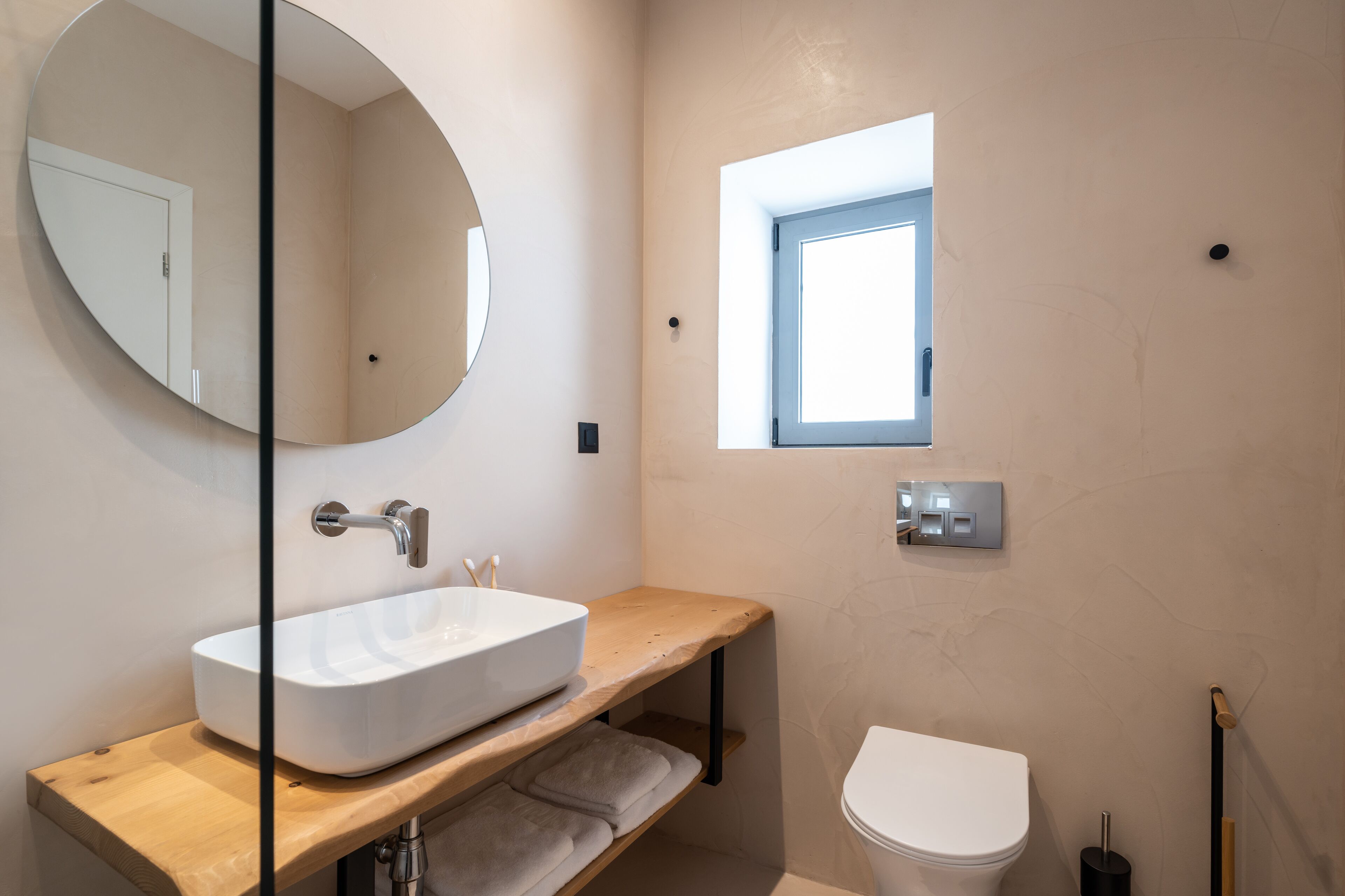 A modern bathroom with a large oval mirror, a white sink, a wooden vanity, a small window, and a toilet.