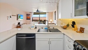 A modern kitchen with a dishwasher, sink, and a black stove. There is a window with a view of the beach and a ceiling fan.