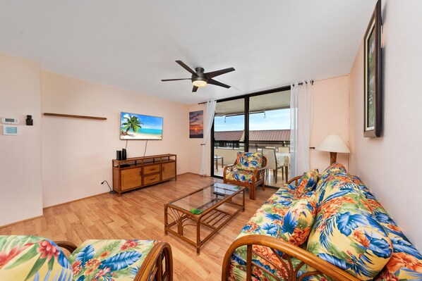 A living room with a wooden floor, a glass coffee table, a sofa with floral upholstery, a ceiling fan, and a view of the outdoors through large windows. - Big Island White Sands Village 323 2 Bedroom Condo (Kailua-Kona)