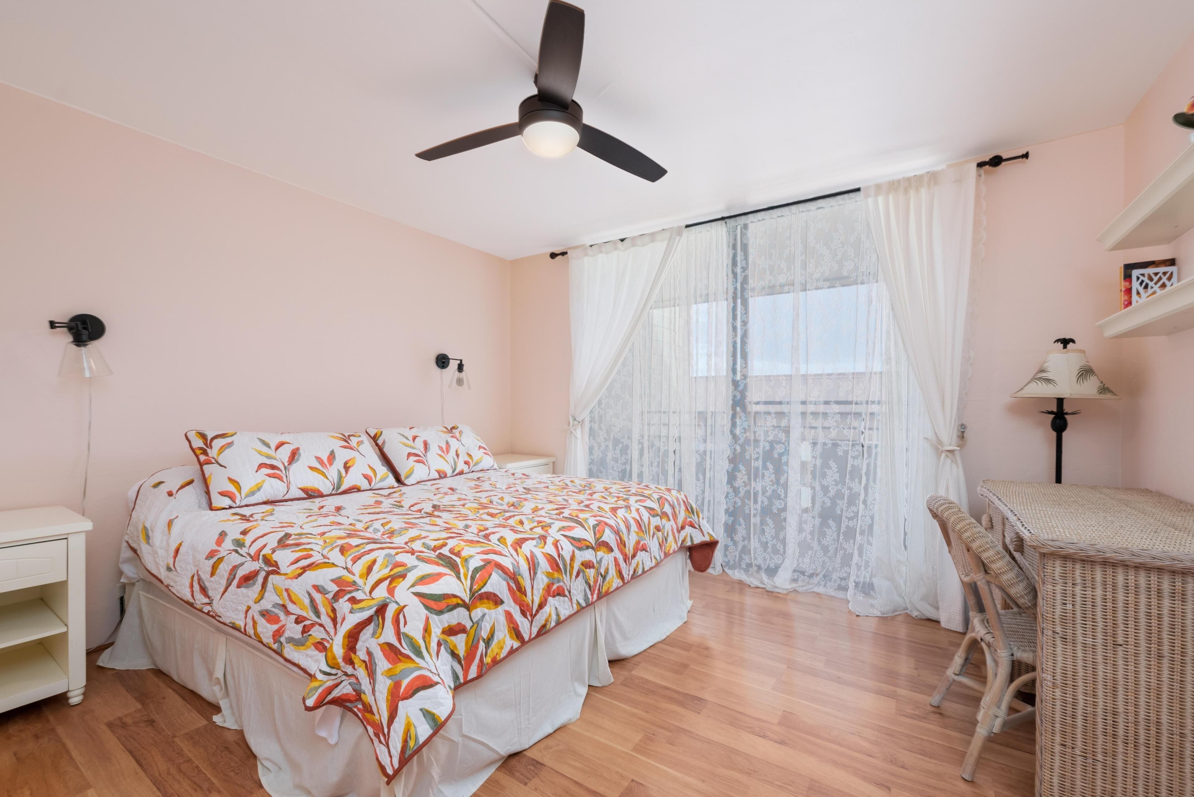 A bedroom with a bed, a ceiling fan, a window with sheer curtains, a bedside table, and a wicker chair.