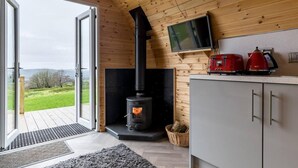 Interior acogedor de una cabaña con estufa de leña, un televisor en la pared y vistas a través de la puerta abierta.