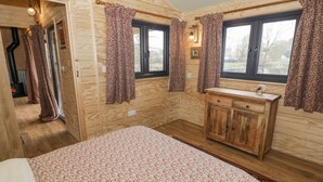 Un dormitorio de cabaña de madera con una cama, un armario de madera con lavamanos y dos ventanas con cortinas estampadas.