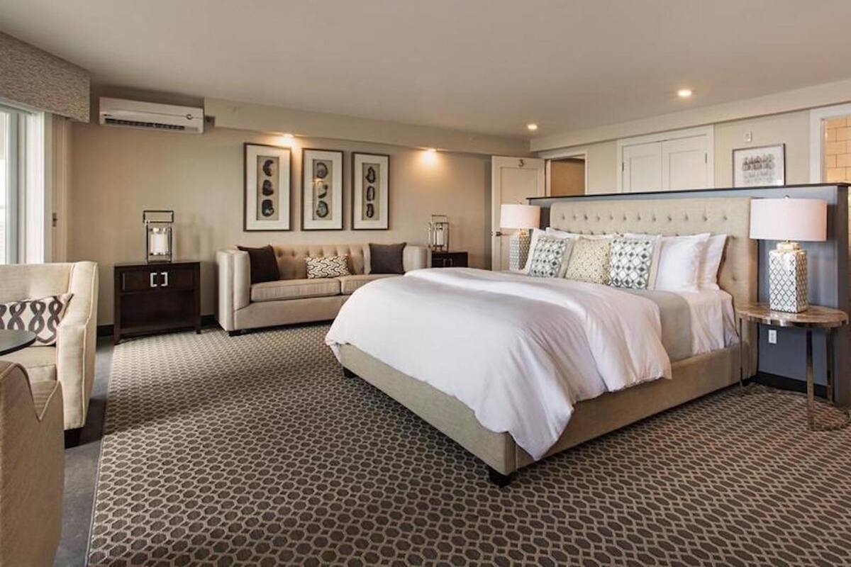 A hotel room with a large bed, a sofa, a small table, and a nightstand. There are framed pictures on the wall and an air conditioning unit above the bed.