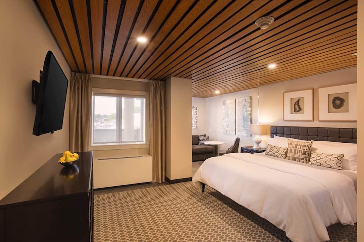 A hotel room with a wooden ceiling, a bed with white bedding, a desk with a chair, a flat-screen TV, and artwork on the walls.