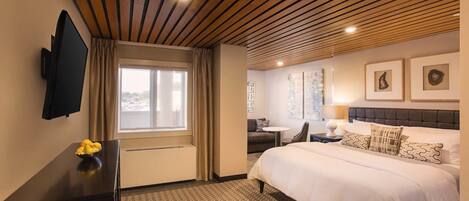 A hotel room with a wooden ceiling, a bed with white bedding, a desk with a chair, a flat-screen TV, and artwork on the walls.