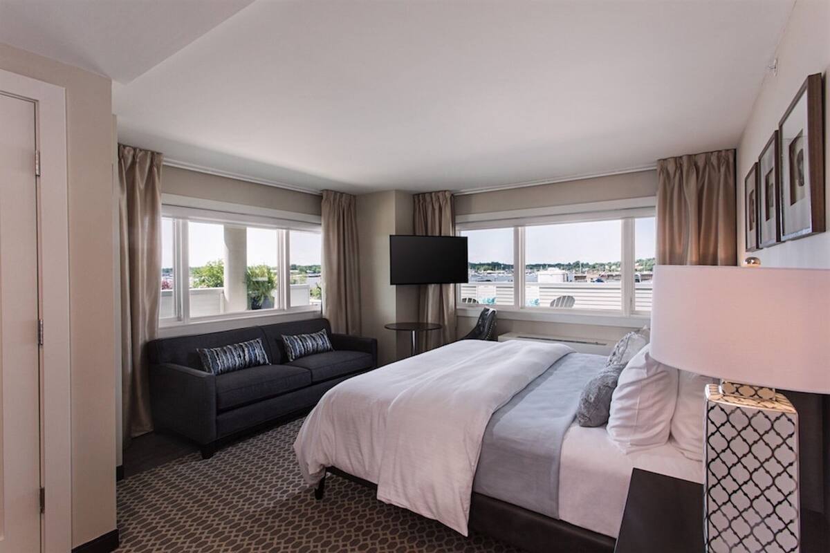A hotel room with a bed, a sofa, a TV, and large windows offering a city view.