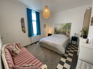 Un dormitorio con cama, sofá, lámpara colgante, ventana con cortinas, un cuadro en la pared y una mesita de noche con lámpara.