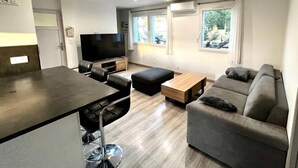 A modern living room with a sofa, ottoman, and a TV. - Cosy apartment, 100 m from beaches, quiet area with outside, air conditioning, wifi & parking (Saint-Chamas)