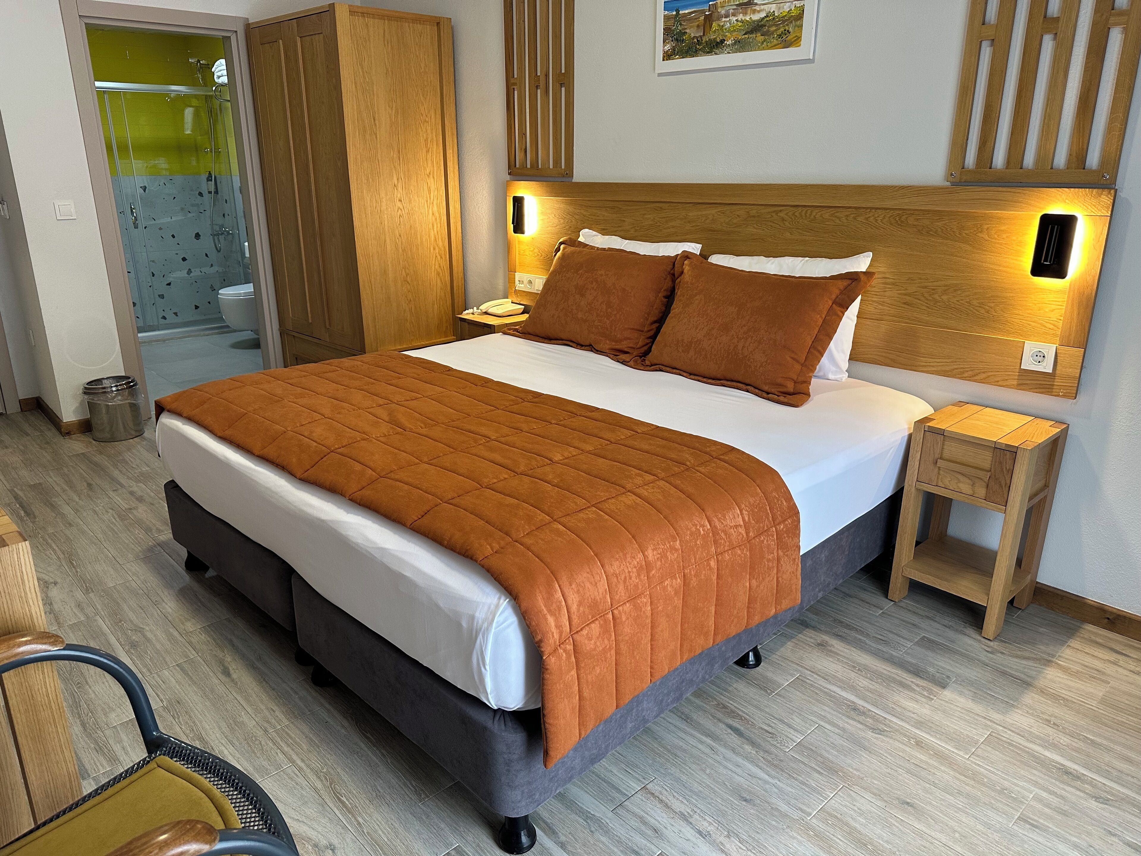 Cama bem arrumada com colcha marrom, cabeceira de madeira e abajures laterais.