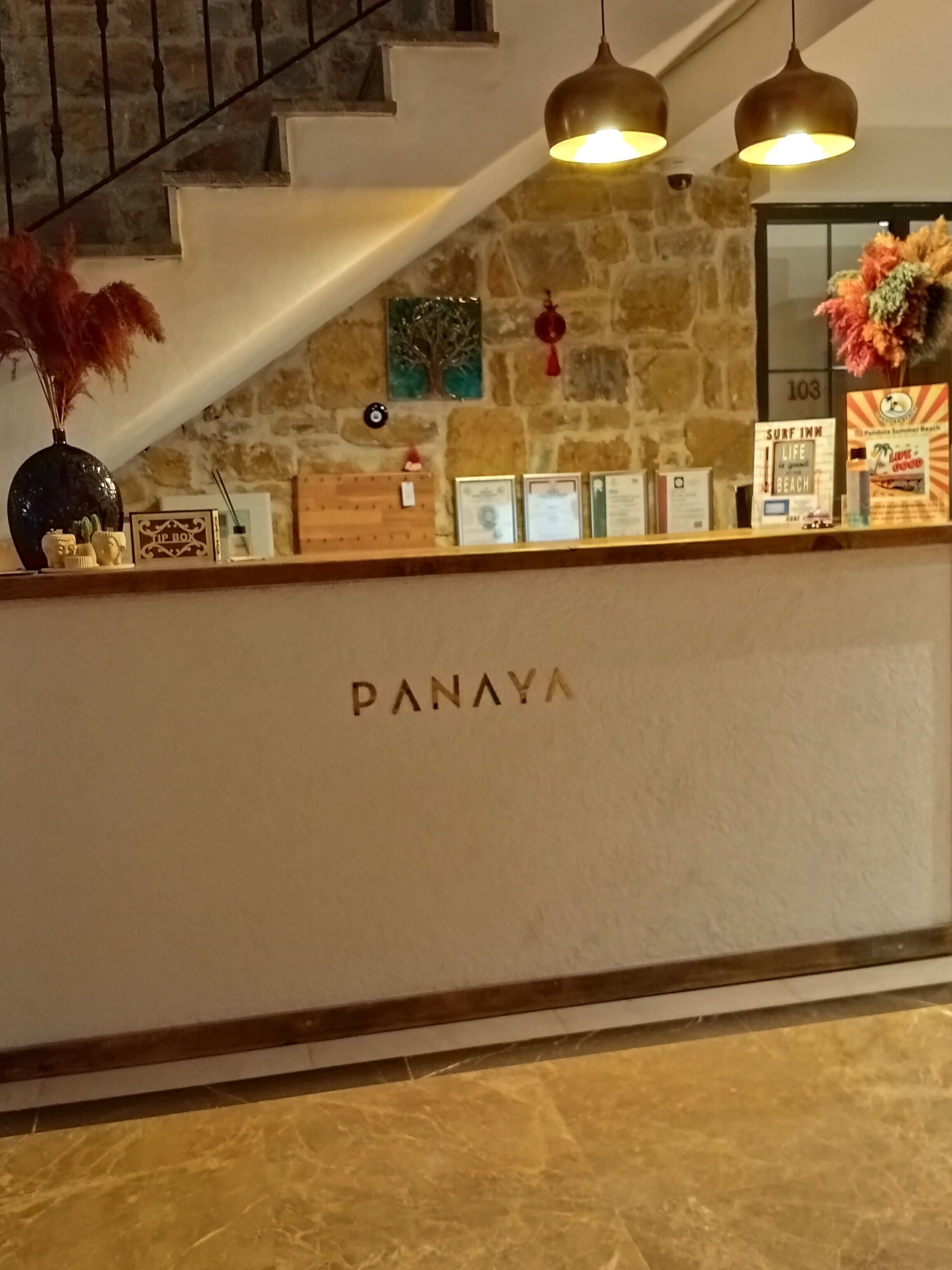A reception desk with the name "PANAYA" in the center, a stone wall, and a staircase in the background.
