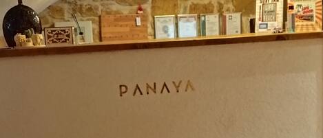 A reception desk with the name "PANAYA" in the center, a stone wall, and a staircase in the background.