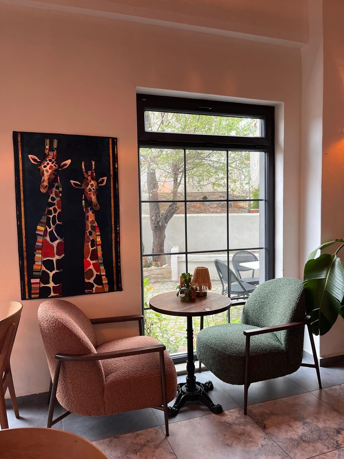 A room with two chairs, a table with a plant, and a giraffe painting on the wall.