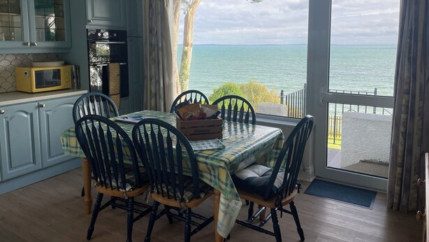 Ein Essbereich mit einem Tisch, gedeckt für vier Personen, umgeben von Stühlen und Blick auf das Meer durch ein großes Fenster.