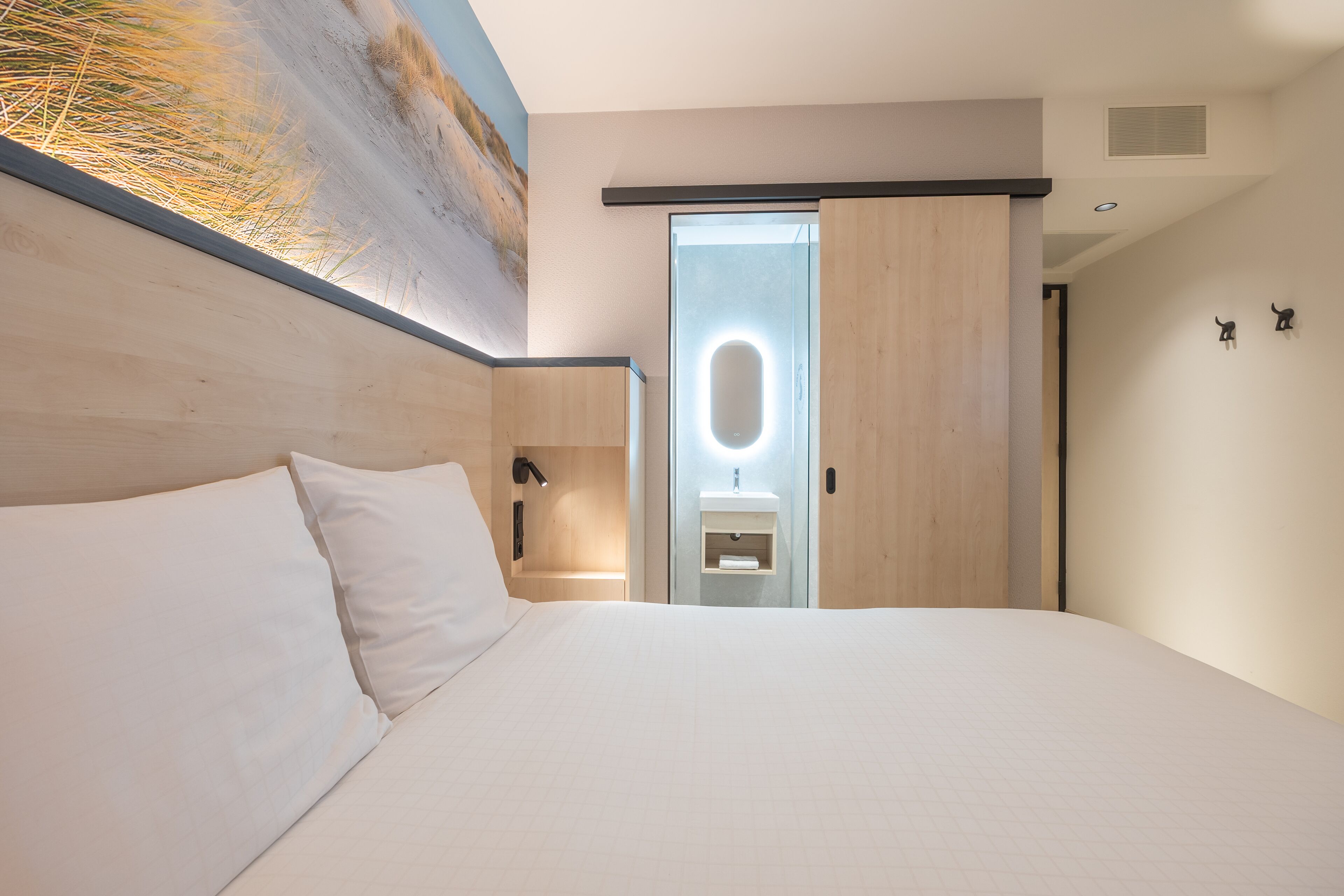 A modern hotel room with a large bed, a wooden headboard, a small bathroom with a mirror, and a painting on the wall.