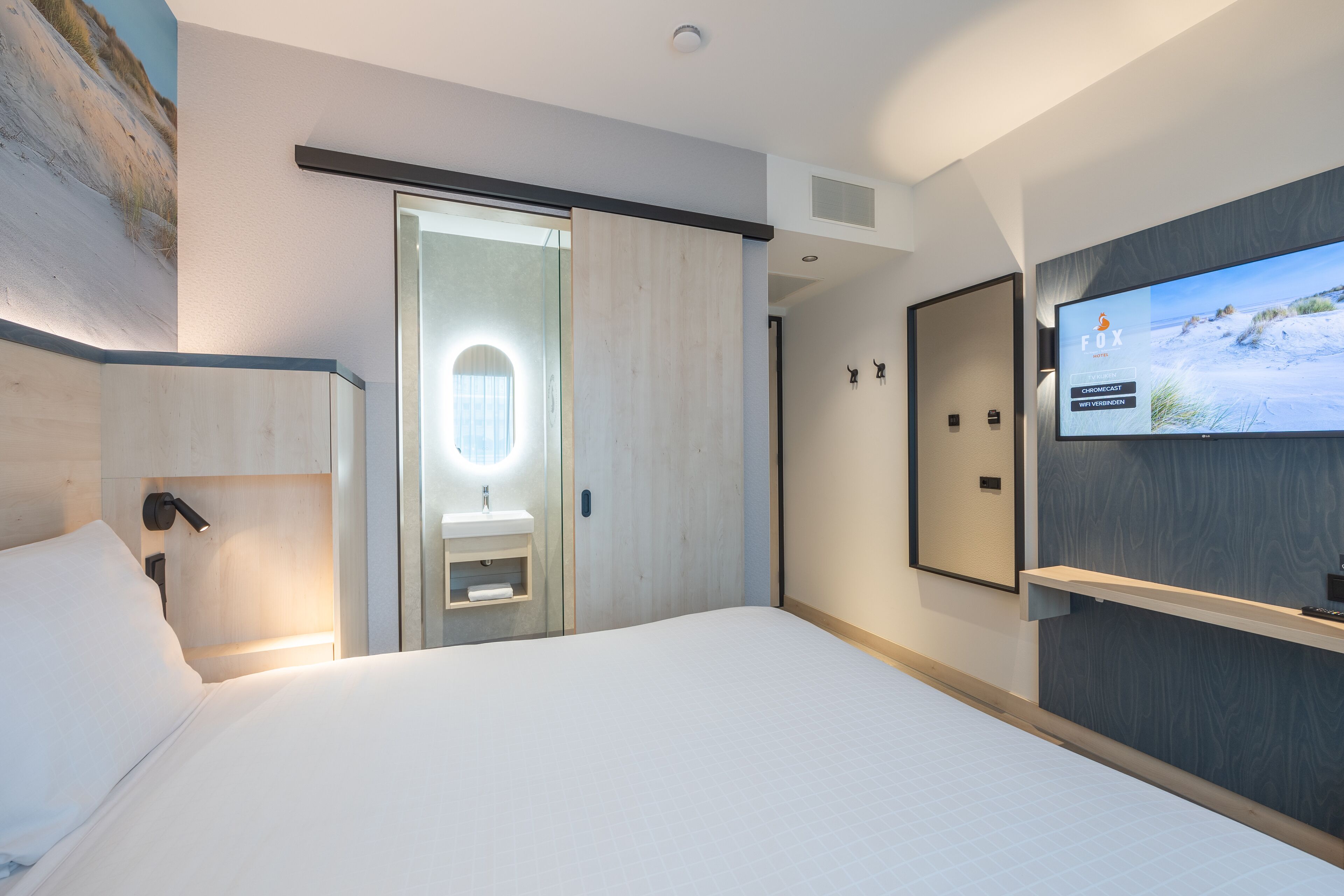 Een moderne hotelkamer met een groot bed, een aan de muur bevestigde televisie, een badkamer met een schuifdeur en een houten hoofdeinde.