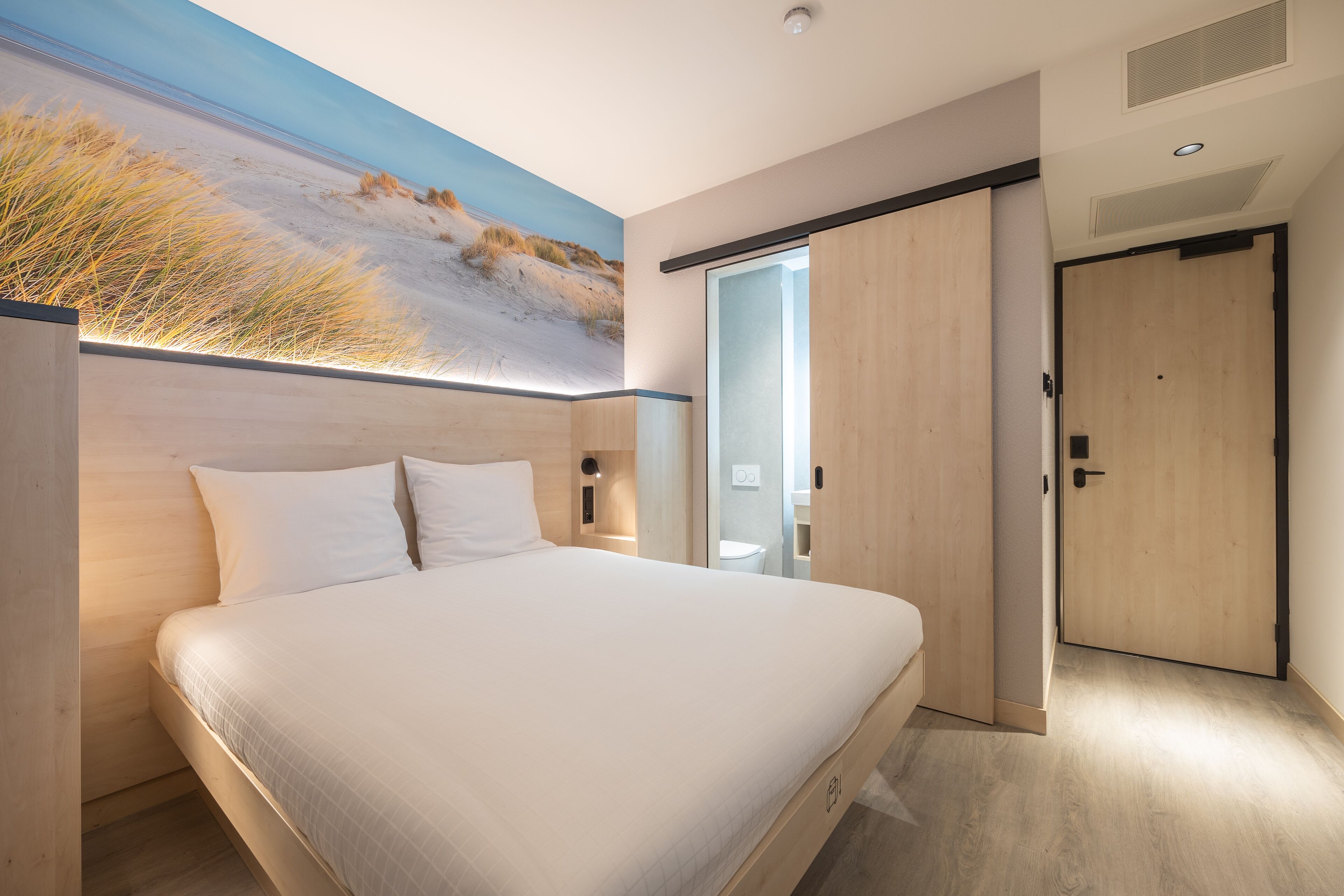 A modern bedroom with a large bed, a painting of a beach scene, and a sliding door leading to a bathroom.