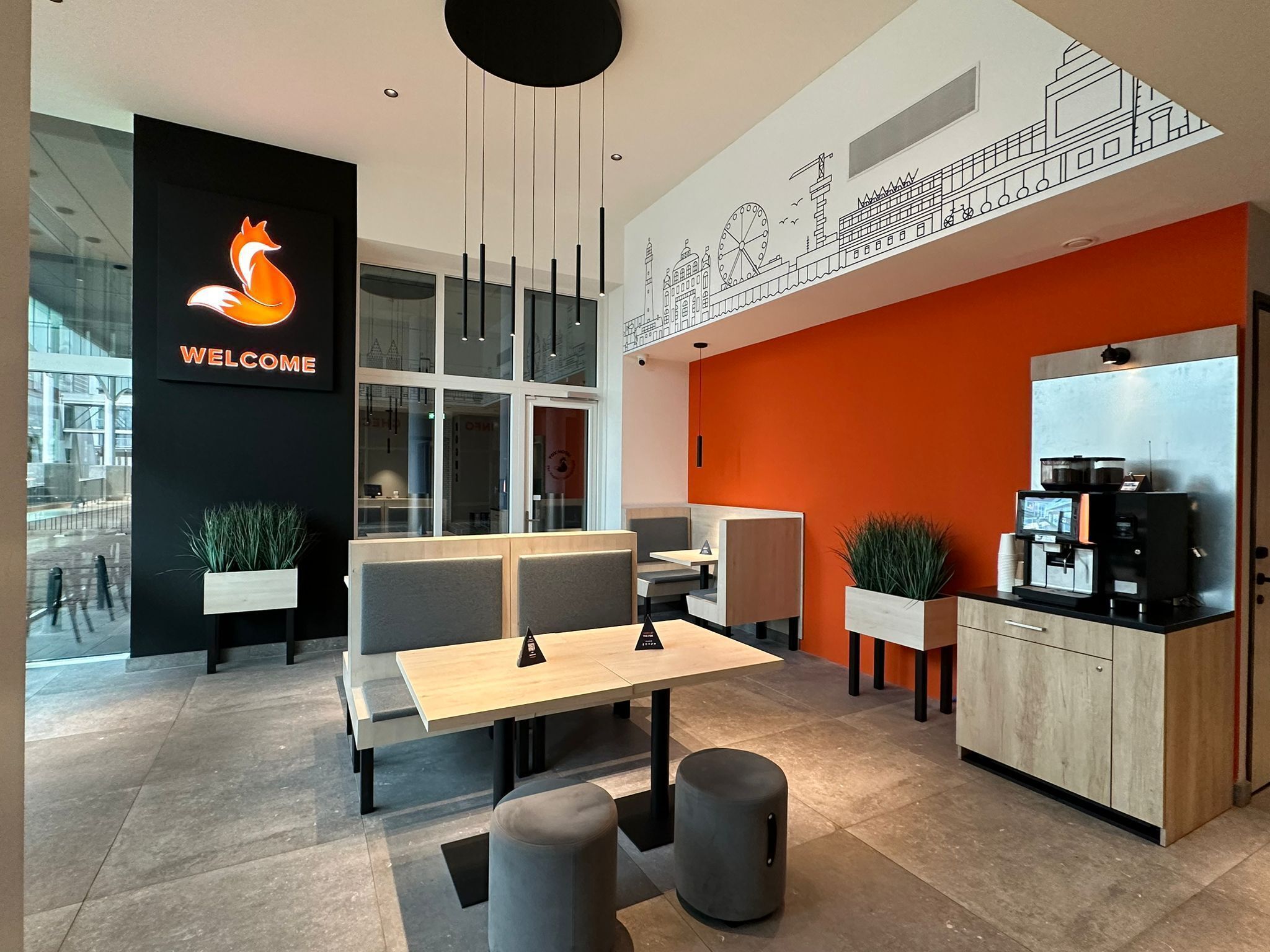 A modern cafe interior with a 'Welcome' sign, seating area, and a coffee machine.