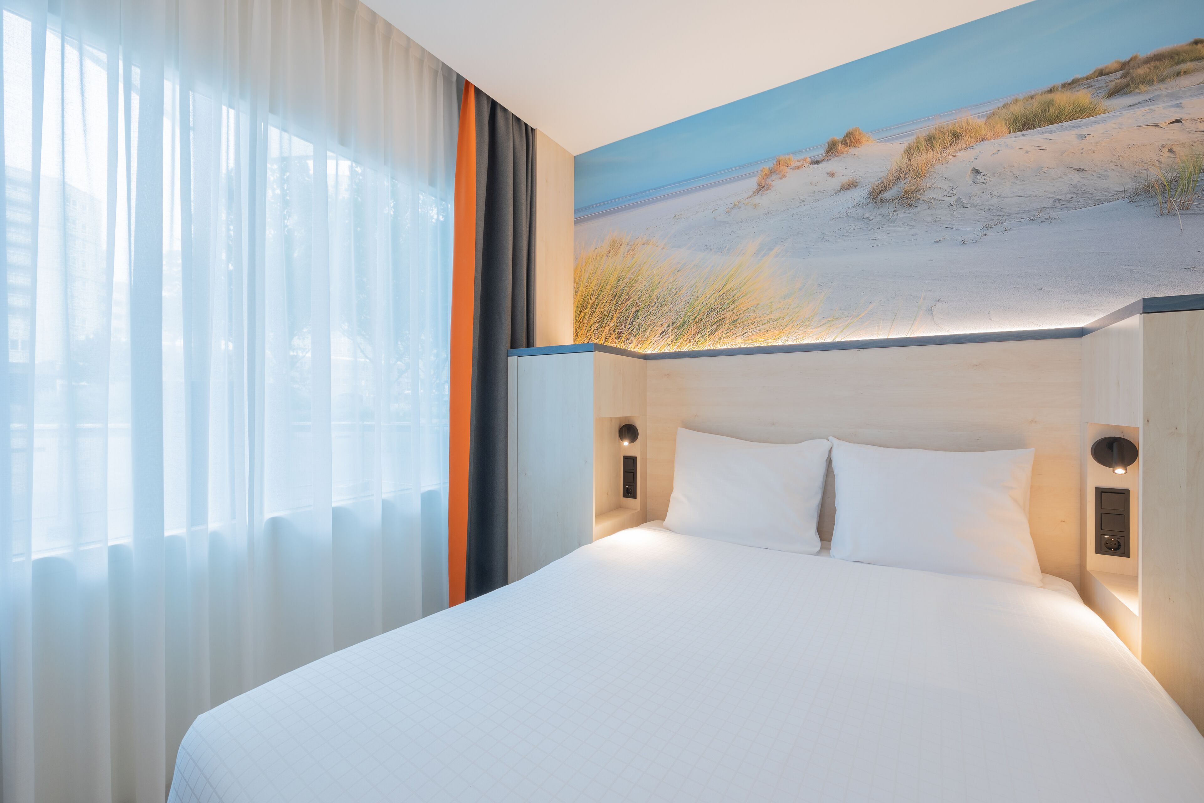 Una habitación de hotel con una cama grande, un cuadro de una playa y una ventana con cortinas traslúcidas.