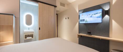 A modern hotel room with a large bed, a TV mounted on the wall, a wooden wardrobe, and a small bedside table.