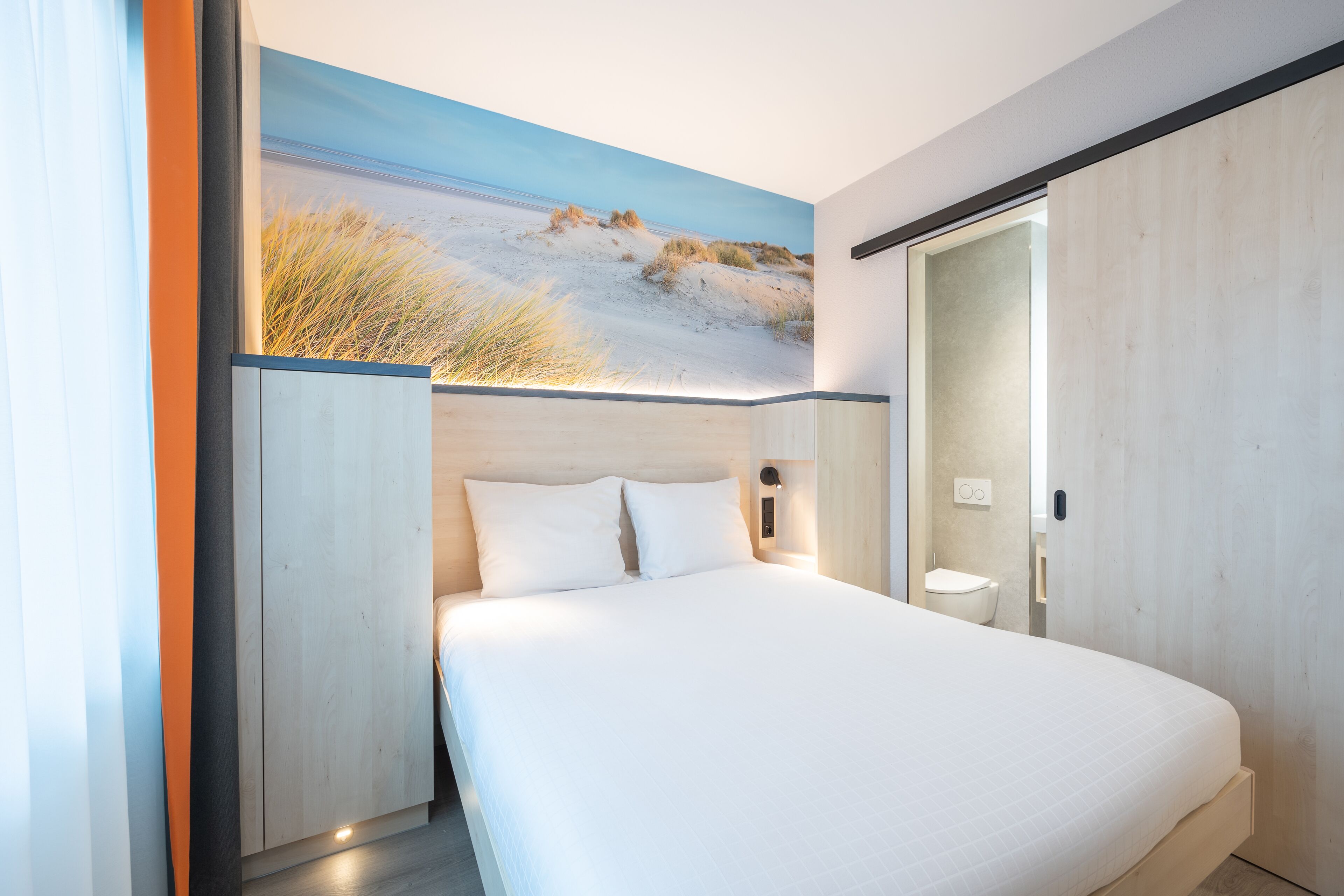 A modern hotel room with a large bed, a wooden wardrobe, and a wall-mounted light fixture.