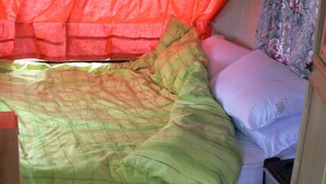 A bed with a green quilt and white pillows, a wooden nightstand, and a red curtain.