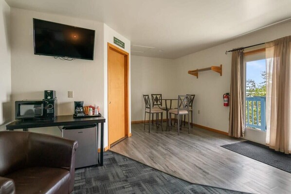 A room with a flat-screen TV, a microwave, a coffee machine, a small refrigerator, a dining table with chairs, and a fire extinguisher.