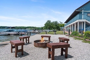 Eine gepflasterte Terrasse mit Feuerstelle und Holztischen, ein Steg mit Booten und ein Gebäude mit blauer Fassade.