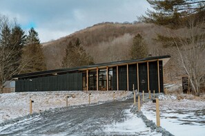 A modern building with a dark facade and large windows, surrounded by a snowy landscape and trees. - Eastwind Oliverea Valley (Big Indian)