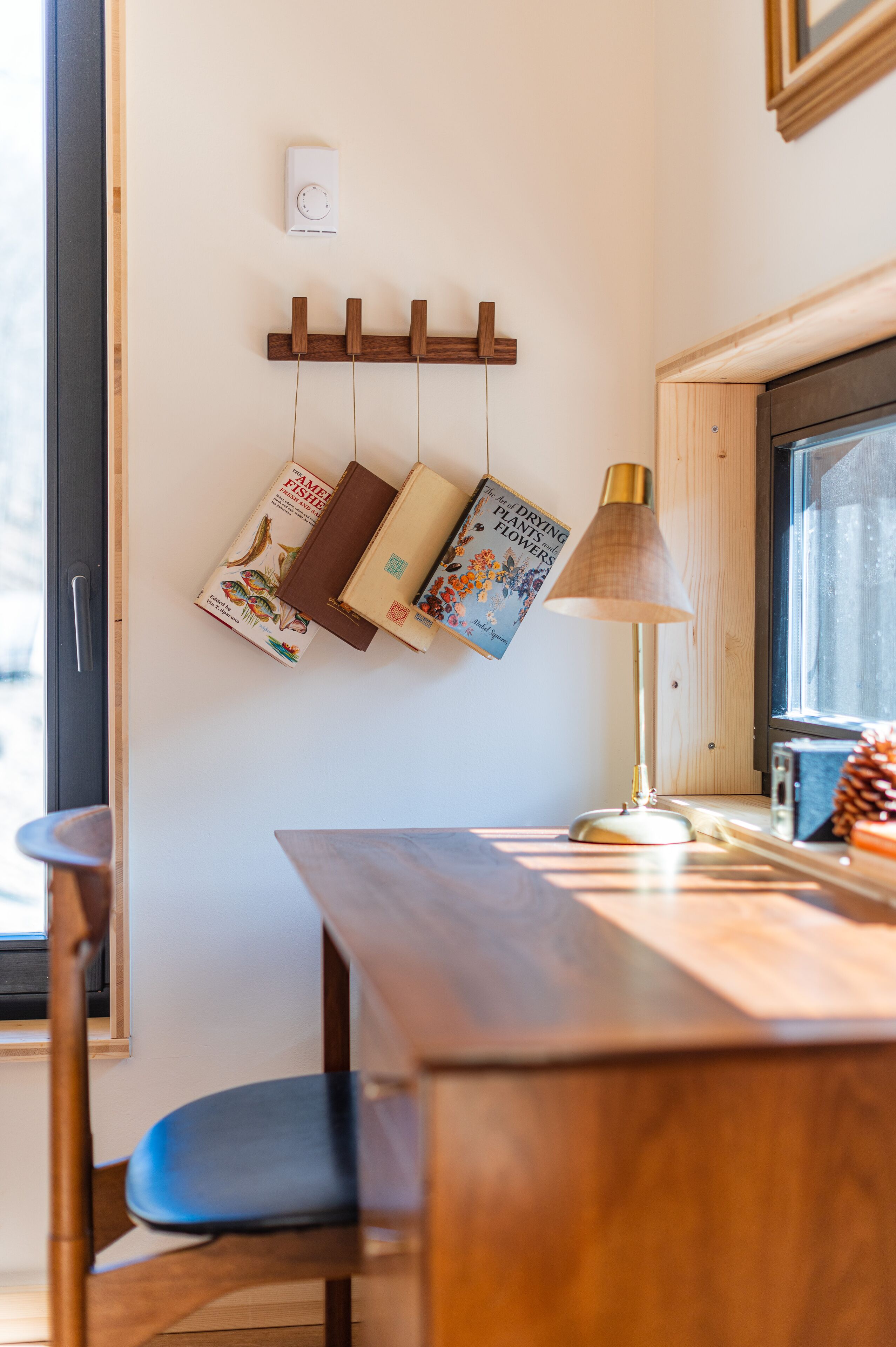 A wooden dining table with a lamp, books on a wall-mounted rack, and a window with a view.